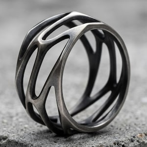 May include: A dark gray metal ring featuring an openwork design with interconnected, branch-like structures. The surface has a matte, brushed finish with darker recessed areas, creating a complex, cage-like appearance throughout the band.