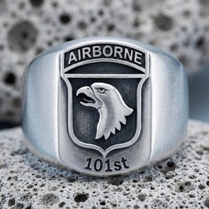 Könnte beinhalten: Ein silberfarbener Metallring mit einem erhabenen schildförmigen Emblem. Das Emblem zeigt oben das Wort AIRBORNE, in der Mitte das Profil eines Adlerkopfes und unten den Text 101st, alles vor einem dunklen, vertieften Hintergrund.