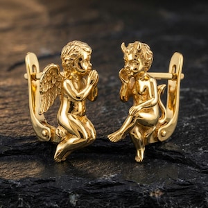 Puede incluir: Un par de pendientes dorados. Uno presenta un ángel arrodillado con alas, el otro un diablo con cuernos y cola. Ambos están en posición sentada, con detalles intrincados.