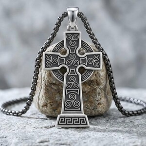 May include: A silver-toned metal Celtic cross pendant featuring intricate spiral triskele engravings and geometric border patterns. The cross hangs from a dark, rounded box chain, showcasing a polished finish with darkened recessed details.
