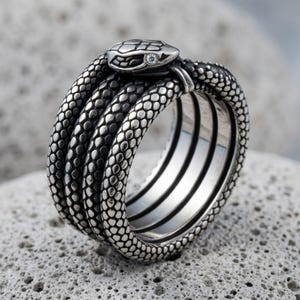 May include: A silver-toned ring shaped like a coiled snake wrapping around the finger multiple times. The metal surface features a detailed, textured scale pattern throughout. A small, clear, round gemstone is set into the snake's head.
