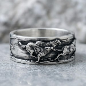 May include: A silver-toned metal ring featuring a detailed, raised relief design of multiple wolves in motion. The band has an oxidised finish that highlights the texture of the running animals against a textured, uneven background surface.