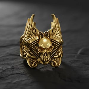 Pode incluir: Um anel dourado com um design de caveira, asas e detalhes do motor. O anel apresenta um design detalhado com uma caveira no centro, ladeada por componentes do motor e encimada por asas estilizadas. O anel está sobre uma superfície escura e texturizada.