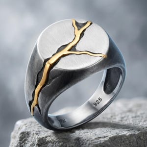 Puede incluir: Un anillo de sello de plata con un acabado oscuro oxidado y una incrustación dorada prominente que parece una grieta ramificada en la cara circular. El interior de la banda está claramente marcado con el número 925.