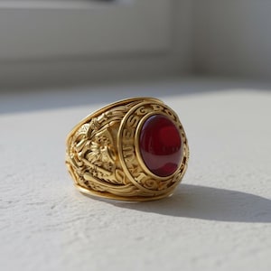 Puede incluir: Un anillo dorado con una gran gema roja ovalada. El anillo tiene detalles intrincados en la banda, incluyendo figuras esculpidas y texto. El anillo está sobre una superficie blanca, con una sombra.