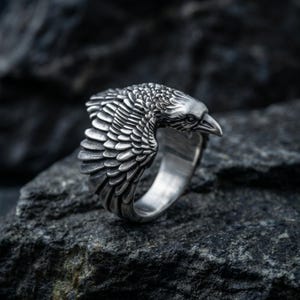 Op de afbeelding: Een gedetailleerde zilveren ring met een gebeeldhouwde raaf met ingewikkelde veerdetaillering. De kop en vleugels van de raaf zijn prominent aanwezig, wat een opvallend accessoire met vogelthema creëert. De ring staat tegen een donkere, getextureerde achtergrond.
