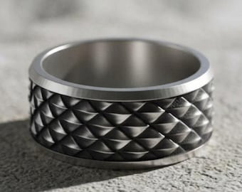 May include: A wide, metallic ring featuring a textured, repeating diamond-shaped pattern across its outer surface. The band has a smooth, polished edge at the top and bottom, with a dark, matte finish within the recessed areas of the geometric design.