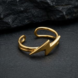 Op de afbeelding: Een goudkleurige ring met een bliksemontwerp. De ring is aan de bovenkant open en heeft een dubbele band. De bliksem is een prominent kenmerk en voegt een gedurfd en stijlvol element toe aan de sieraden.