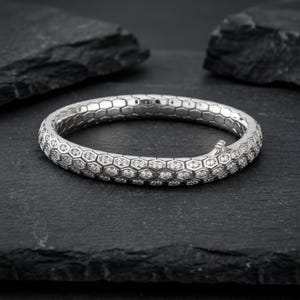 Op de afbeelding: Een zilveren armband met een honingraatontwerp, bezet met kleine, sprankelende edelstenen. De armband wordt gepresenteerd op een donkere, getextureerde achtergrond, die de ingewikkelde details en elegante uitstraling benadrukt.