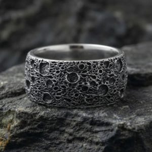Puede incluir: Un anillo plateado con una superficie texturizada que se asemeja a la superficie lunar, con numerosos cráteres de varios tamaños. El anillo se encuentra sobre un fondo oscuro y texturizado, resaltando su diseño detallado. Este anillo es una joya única.