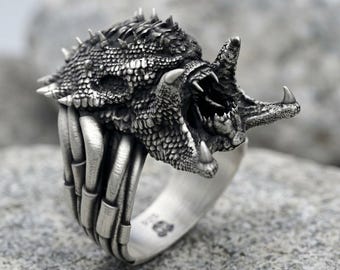 May include: A silver ring featuring a detailed, textured creature head with sharp spikes, an open mouth revealing teeth, and a band designed to resemble a series of connected bone-like segments. The metal has an oxidized finish to highlight details.