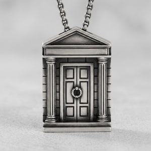 May include: A silver-toned metal pendant shaped like a miniature temple facade with a pediment roof, two fluted columns, and a central double door featuring a circular knocker. The pendant hangs from a matching silver-toned metal chain link necklace.