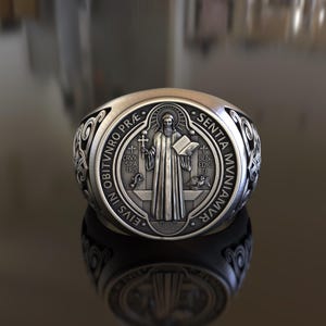 Puede incluir: Un anillo de plata con un diseño detallado que presenta una figura religiosa y la inscripción "EIVS IN OBITVNRO PRÆ. SENTIA MUNIAMVR".