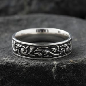May include: A silver ring with an intricate floral pattern. The ring's band features a detailed design of swirling leaves and vines, creating a textured appearance. The ring is set against a dark, textured background, highlighting its metallic sheen.