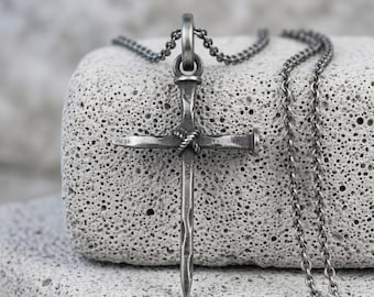 May include: A dark silver-toned metal pendant shaped like a cross, constructed from two intersecting nail-like pieces bound together at the center with a twisted wire detail. The pendant hangs from a matching thin, delicate metal link chain.