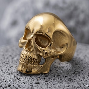 Puede incluir: Un anillo de tono dorado pulido con forma de cráneo humano. El anillo presenta una superficie lisa y altamente reflectante con hendiduras detalladas para las cuencas de los ojos, la cavidad nasal y los dientes, capturando una estructura anatómica.