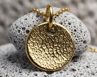 May include: A circular gold pendant featuring a highly textured, organic surface with deep, irregular indentations. The pendant is suspended from a delicate gold chain by a smooth, vertical loop bail, creating a contrast between the rough metal finish.