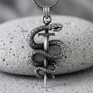 May include: A silver-toned metal pendant featuring a detailed snake coiled around a vertical sword. The snake has visible scales and an open mouth, while the sword has a simple crossguard and a pointed tip. The pendant hangs from a silver chain.