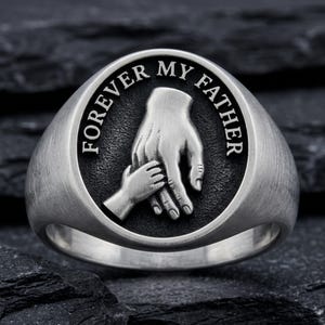 May include: A silver-colored ring with the words "FOREVER MY FATHER" engraved around a central design. The design features two hands, one adult and one child, holding each other. The ring has a smooth, polished finish.
