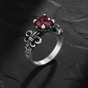 Puede incluir: Un anillo plateado con una gran gema roja redonda. El anillo presenta un diseño de flor de lis en la banda y detalles intrincados alrededor del engaste de la gema. El anillo se muestra sobre una superficie oscura y texturizada.