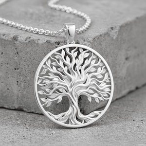 May include: A circular silver pendant featuring an intricate tree of life design with detailed branches, leaves, and roots, suspended from a matching silver chain. The metallic surface has a polished finish that reflects light across the relief.