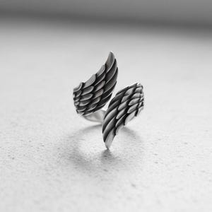 May include: A silver ring with an open wing design. The ring's wings are detailed, with each feather clearly defined. The ring is set against a neutral background, emphasising the intricate craftsmanship and the ring's metallic sheen.