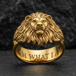 May include: A gold-colored ring featuring a detailed lion's head design. The ring has the engraved text "I AM WHAT I AM" on the band. The lion's mane is intricately sculpted, and the ring has a polished finish.