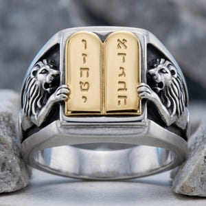 May include: A silver-toned ring featuring a central gold-toned rectangular plaque engraved with Hebrew characters. The plaque is flanked by two sculpted lion figures on either side, with a dark, oxidized finish highlighting the intricate details.