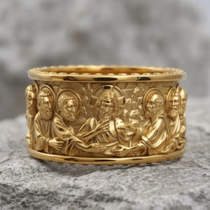 Pode incluir: Um anel de banda larga em ouro com um relevo detalhado da Última Ceia. O design mostra uma figura central com auréola rodeada por vários indivíduos barbudos com túnicas, tudo sobre uma superfície de fundo texturizada e irregular.