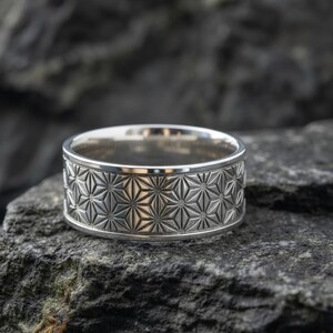 Könnte beinhalten: Ein silberner Ring mit einem geometrischen Sternmuster. Der Ring ist breit und liegt auf einer dunkelgrauen Steinoberfläche. Das Design des Rings zeigt sich wiederholende Sternformen um das Band.