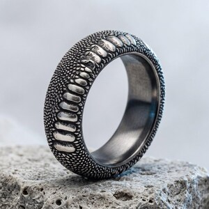 May include: A silver-toned metal ring featuring a textured surface that mimics the appearance of reptile skin, with a row of raised, elongated scales running along the centre and a smooth, dark interior band finish.