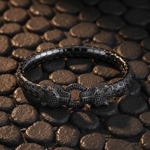 Könnte beinhalten: Ein dunkelgraues Armband mit einem Pantherkopf-Design. Das Armband hat zwei Pantherköpfe, die sich gegenüberliegen und mit schwarzen Steinen besetzt sind. Das Armband hat ein sechseckiges Muster.