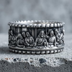Pode incluir: Um anel largo de metal prateado com um relevo tridimensional detalhado da cena da Última Ceia. A banda é contornada por um design de borda festonada, exibindo figuras sentadas a uma mesa longa com taças e pratos dispostos.