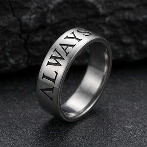 May include: A silver ring with the word "ALWAYS" engraved in black capital letters. The ring has a brushed metal finish and a simple, classic design. The ring is shown on a dark, textured surface.