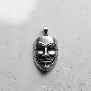 May include: A silver-colored pendant in the shape of a stylized mask, featuring a smiling face with a mustache and goatee. The pendant has a loop at the top for a chain. The background is a textured, off-white surface.