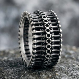 May include: A silver-toned metal ring featuring a repeating pattern of vertical bars and a central row of small, circular cutouts. The surface has a darkened, oxidized finish in the recessed areas, highlighting the structural details of the band.
