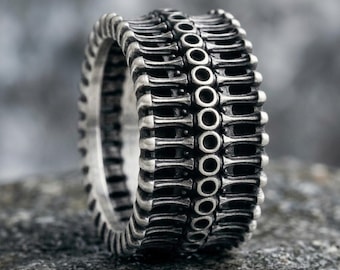 May include: A silver-toned metal ring featuring a repeating pattern of vertical bars and a central row of small, circular cutouts. The surface has a darkened, oxidized finish in the recessed areas, highlighting the structural details of the band.
