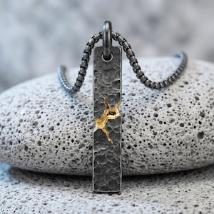 May include: A rectangular pendant with a dark, hammered, charcoal-coloured finish featuring a jagged, gold-coloured fissure running across the centre. The pendant hangs from a matching dark, rounded box chain, creating a high-contrast metallic look.