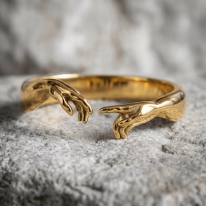 May include: A gold-toned ring featuring two sculpted hands reaching toward each other. The band is open at the front, with each end shaped like a hand with fingers extended. The metal has a polished, reflective surface that catches the light.