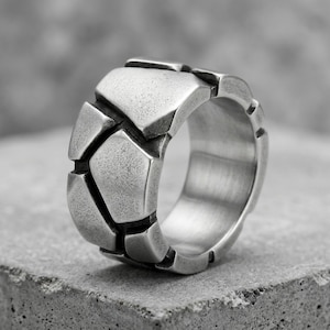 May include: A wide silver-toned metal ring featuring a textured surface composed of multiple irregular, geometric, stone-like segments separated by deep, dark grooves. The band has a smooth, polished interior and a matte, weathered exterior finish.