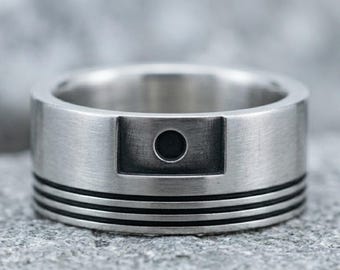 May include: A wide silver-toned metal ring with a brushed finish. The band features a rectangular recessed section containing a single circular black inlay. Three thin, horizontal black grooves are engraved around the lower portion of the ring.