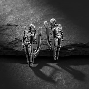 May include: A pair of silver-toned robot-shaped earrings. Each earring features a detailed humanoid robot design with intricate mechanical detailing. The earrings are shown against a dark, textured background, highlighting their metallic finish.
