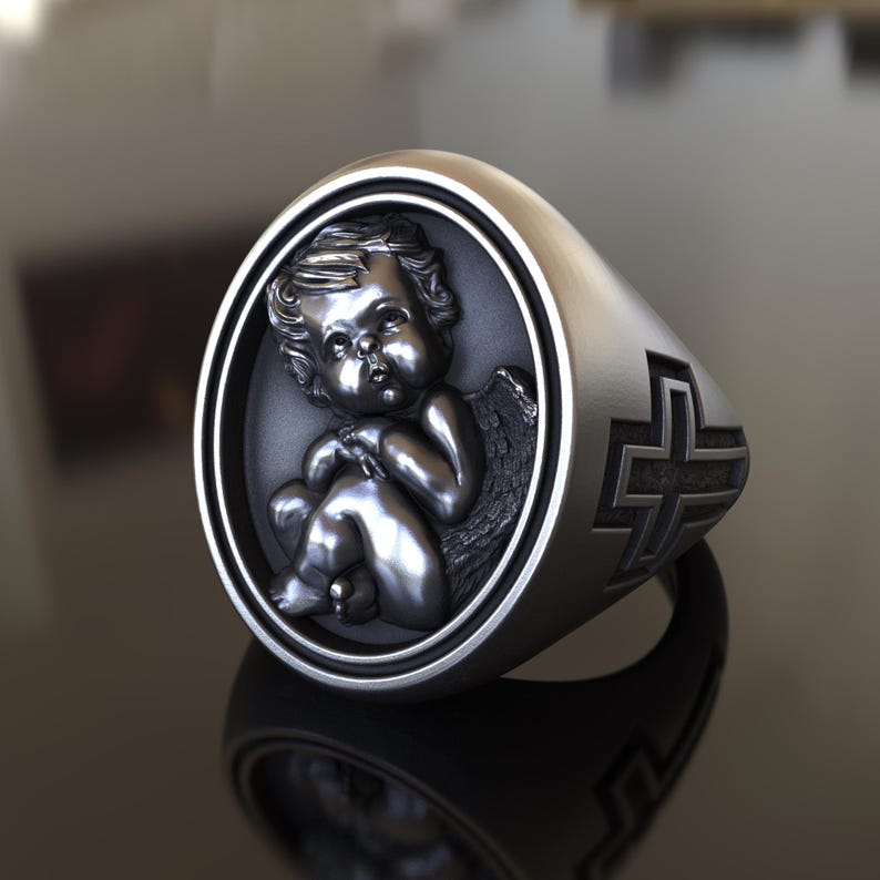 May include: A silver ring with a detailed cherub design in a circular setting and a cross design on the band.