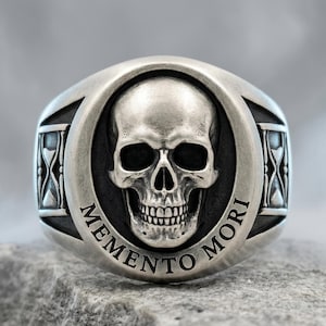 May include: A silver-toned ring featuring a central raised human skull motif set against a dark recessed background. The words MEMENTO MORI are engraved along the lower edge of the circular frame, with stylized hourglass symbols on the sides.