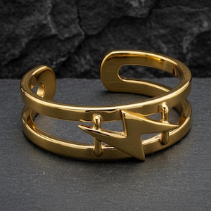 Op de afbeelding: Een goudkleurige manchetarmband met een bliksemontwerp. De open manchet heeft een dubbelbandig ontwerp met een bliksemschicht in het midden. De armband is gemaakt van een glanzend, metallic materiaal en wordt gepresenteerd op een donkergrijze ondergrond.