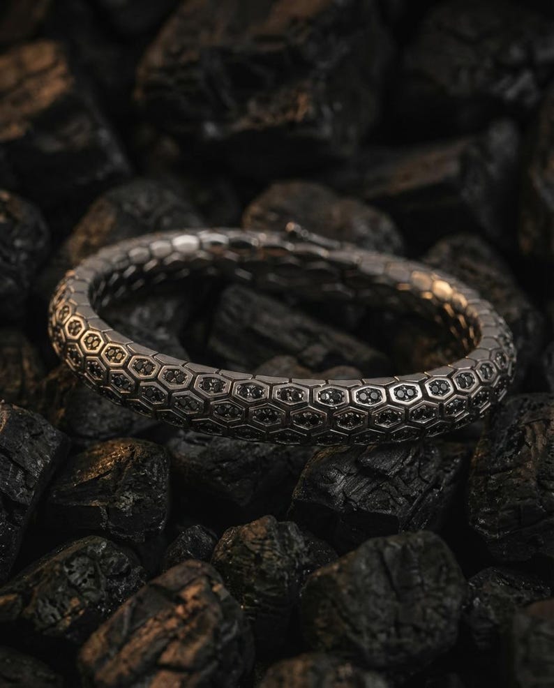 Puede incluir: Una pulsera plateada con un dise&ntilde;o hexagonal y detalles negros. La pulsera descansa sobre un lecho de piedras oscuras y texturizadas. El dise&ntilde;o presenta un patr&oacute;n repetitivo de peque&ntilde;as piedras oscuras dentro de cada celda hexagonal.