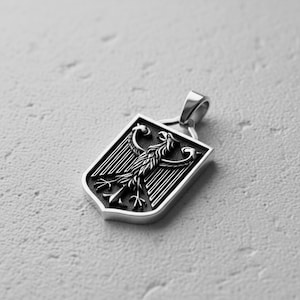 May include: A silver-toned metal pendant shaped like a shield, featuring a detailed embossed eagle emblem with outstretched wings against a dark, recessed background. The pendant includes a polished bail at the top for attachment to a chain.