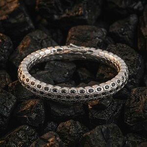 Puede incluir: Un brazalete plateado con dise&ntilde;o de panal, con peque&ntilde;as piedras negras engastadas en cada celda. El brazalete se exhibe sobre un lecho de carb&oacute;n vegetal oscuro y texturizado, creando un contraste entre el metal brillante y el fondo rugoso.