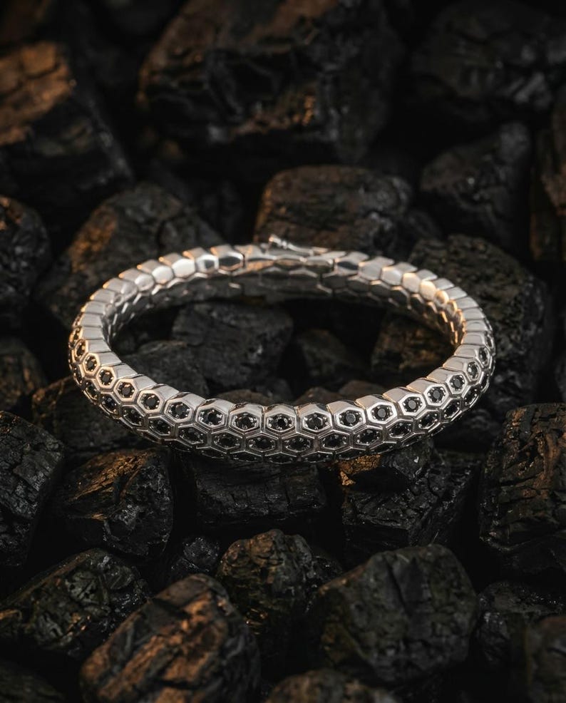 Puede incluir: Un brazalete plateado con dise&ntilde;o de panal, con peque&ntilde;as piedras negras engastadas en cada celda. El brazalete se exhibe sobre un lecho de carb&oacute;n vegetal oscuro y texturizado, creando un contraste entre el metal brillante y el fondo rugoso.