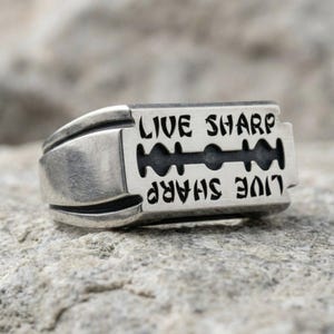 May include: A silver-toned metal ring shaped like a razor blade. The surface features a recessed black cutout design resembling a blade slot. The text "LIVE SHARP" is engraved twice on the front, with one instance appearing in an inverted orientation.